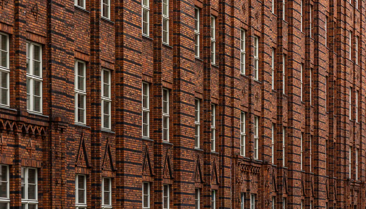 Rotbraune Klinkerfassade eines mehrgeschossigen Gebäudes mit dunklen Klinkerbändern, weißen Sprossenfenstern und dreieckigen Zierelementen aus Klinker